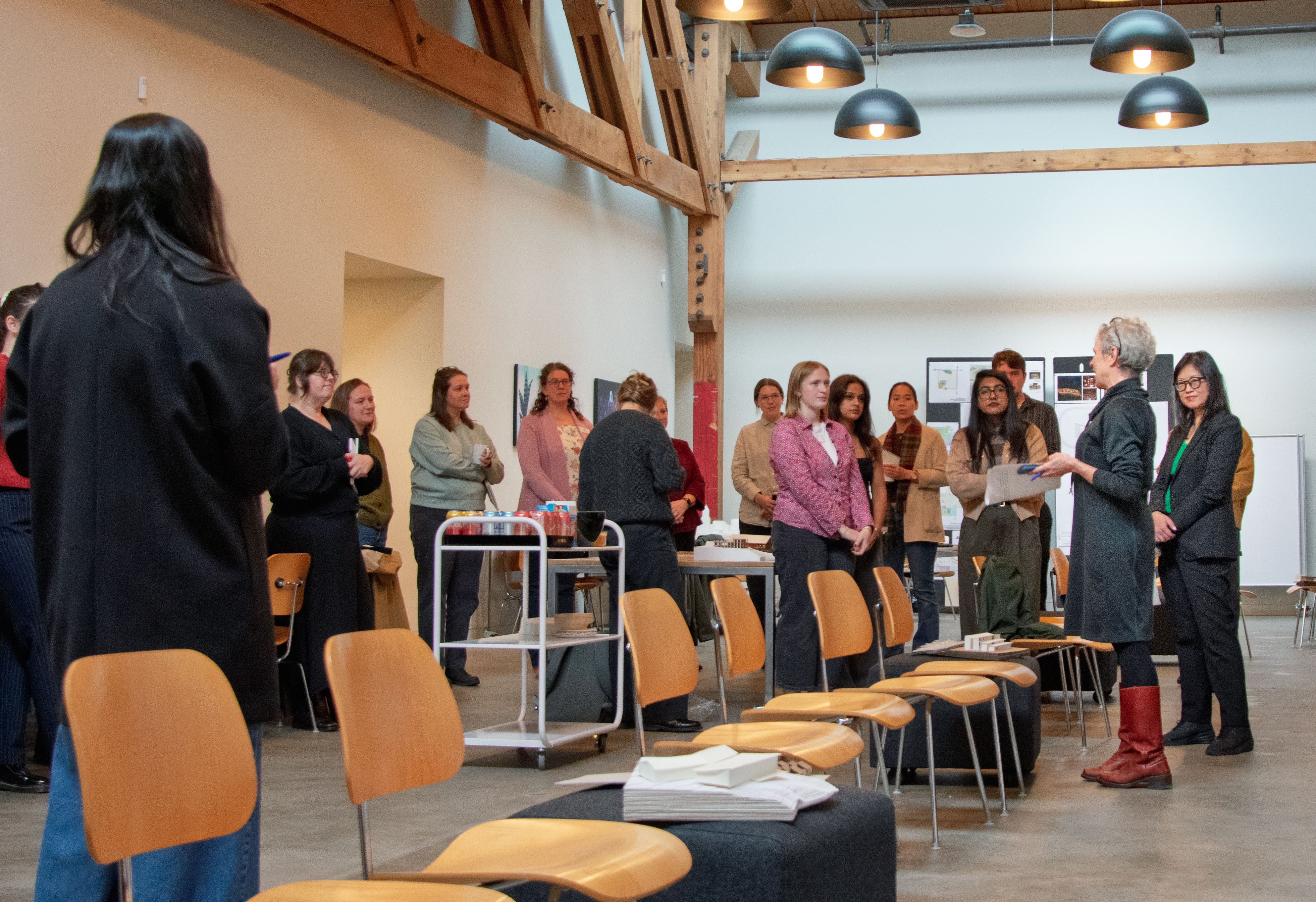 A large room with wooden beams, students listen to two speakers at the right. Chairs are et up in front of large posters. 