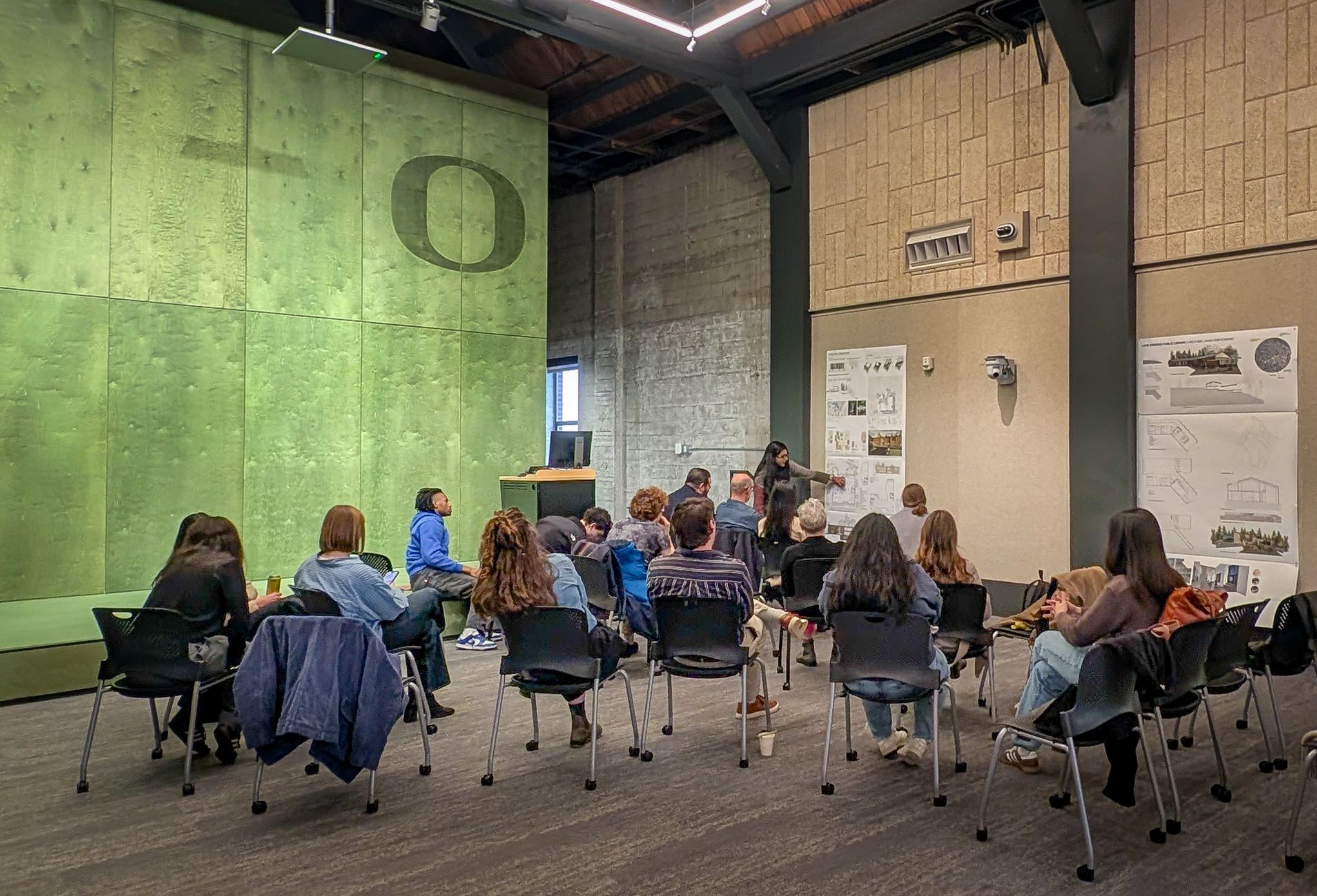 A UO Portland architecture student describes the final project for the library design studio. The students are in the Innovation Common, with a bright green wall and the "O".