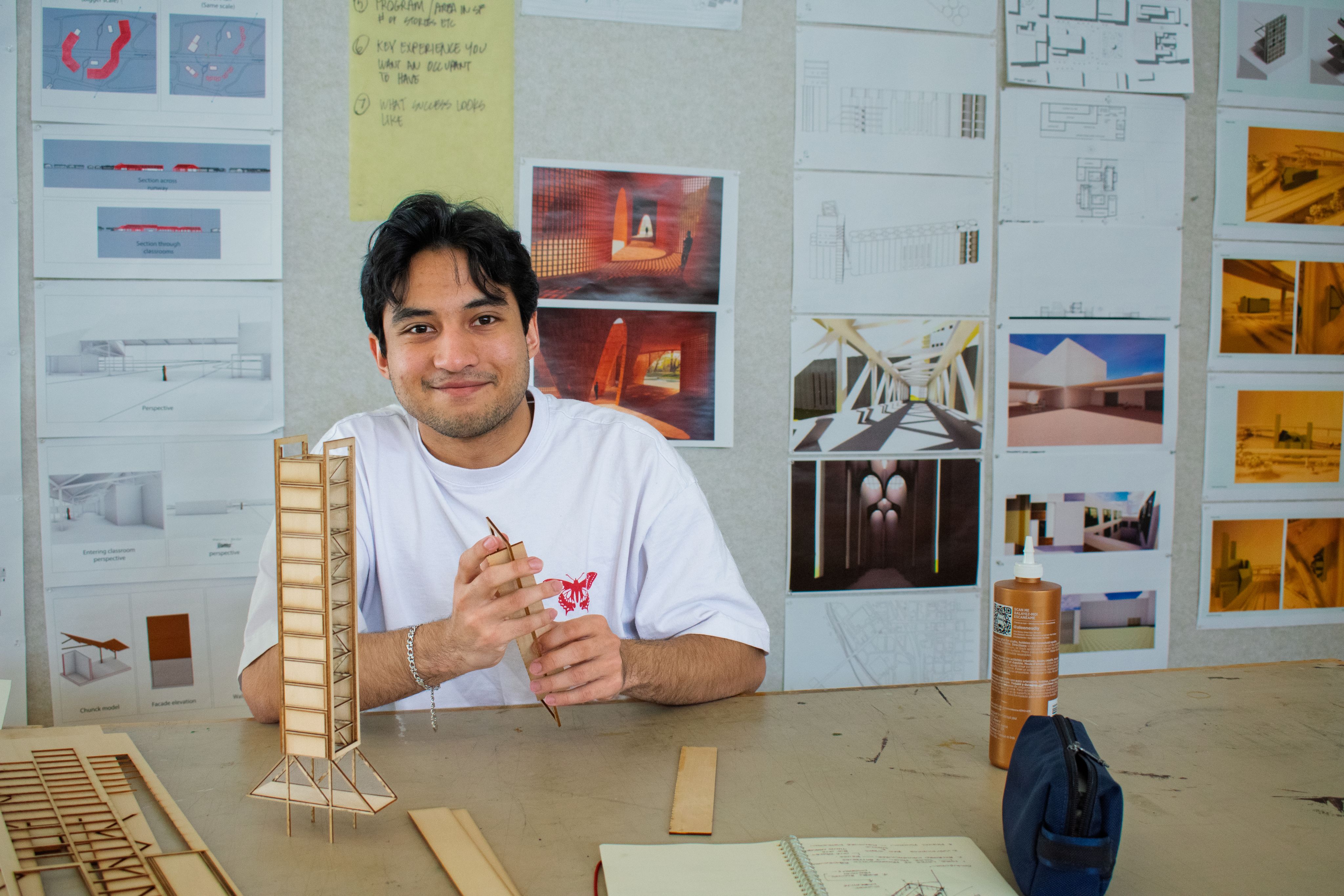 Gabriel Alvarado sits at a table working on architecture models. A sketch book is in front of him and architectural renderings on the wall behind. 