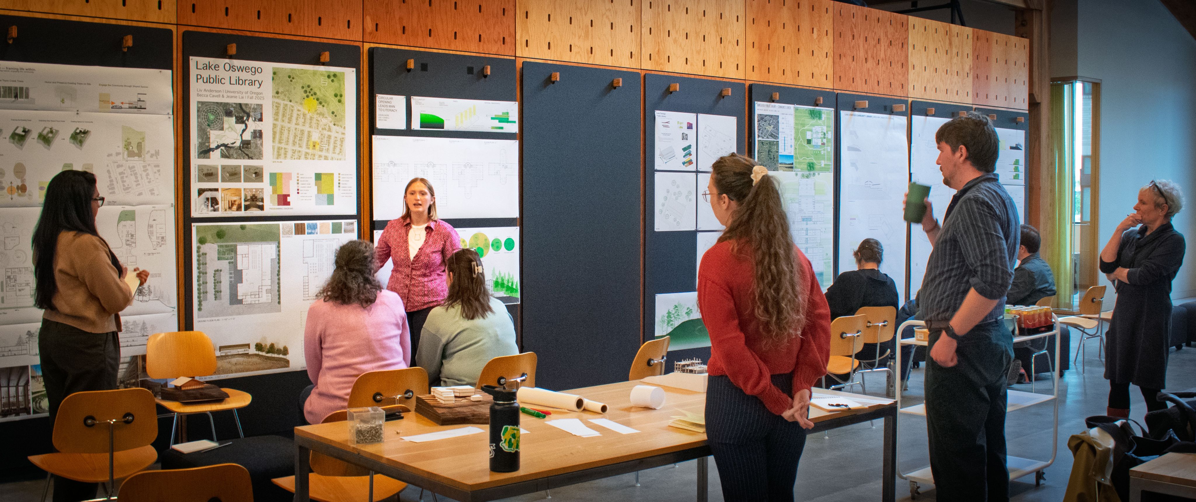 Students discuss projects in the Bora office. Six posters along the back wall. Audience sitting and standing.