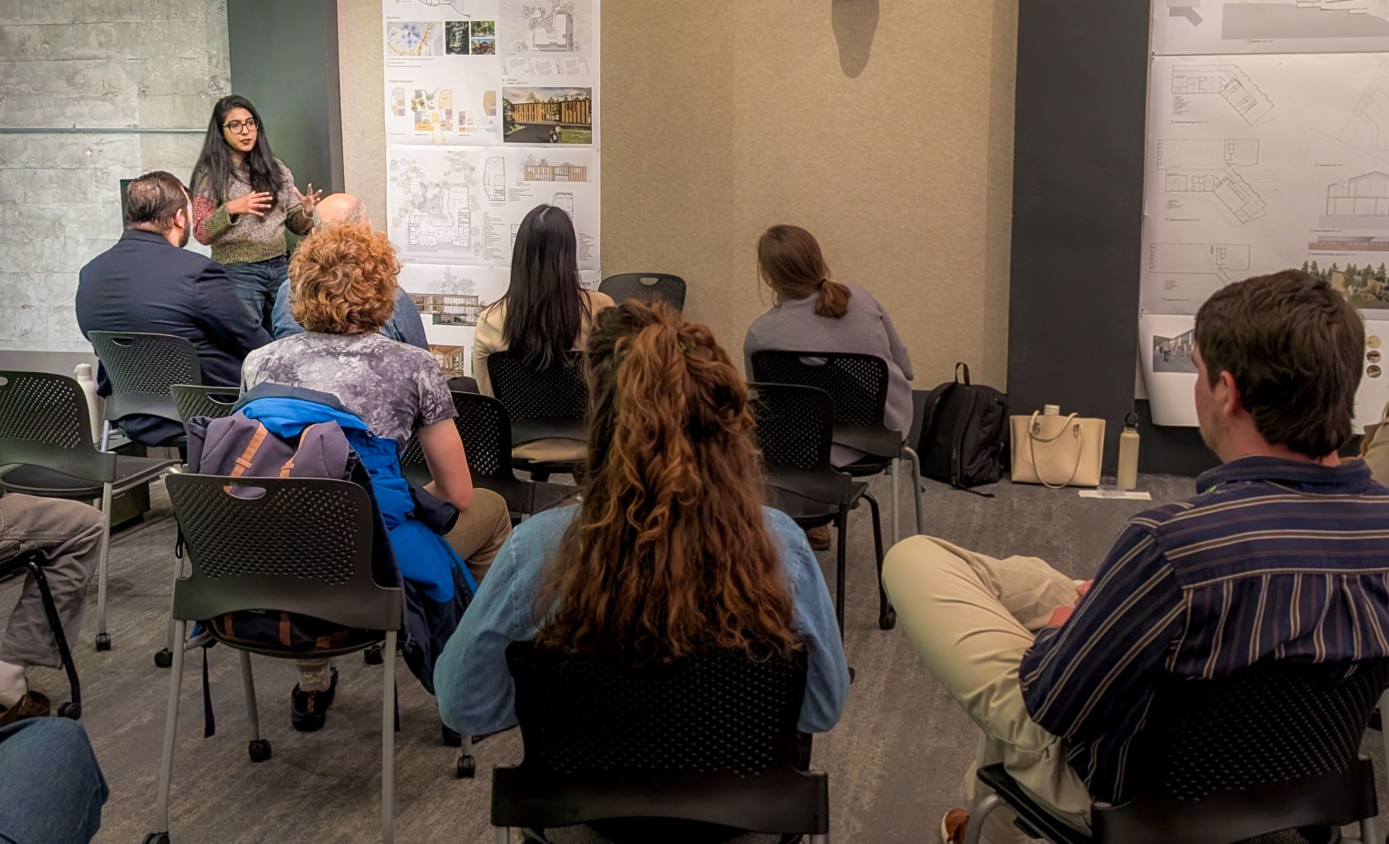 A student stands near a poster at a final review with about 10 people seated in the audience
