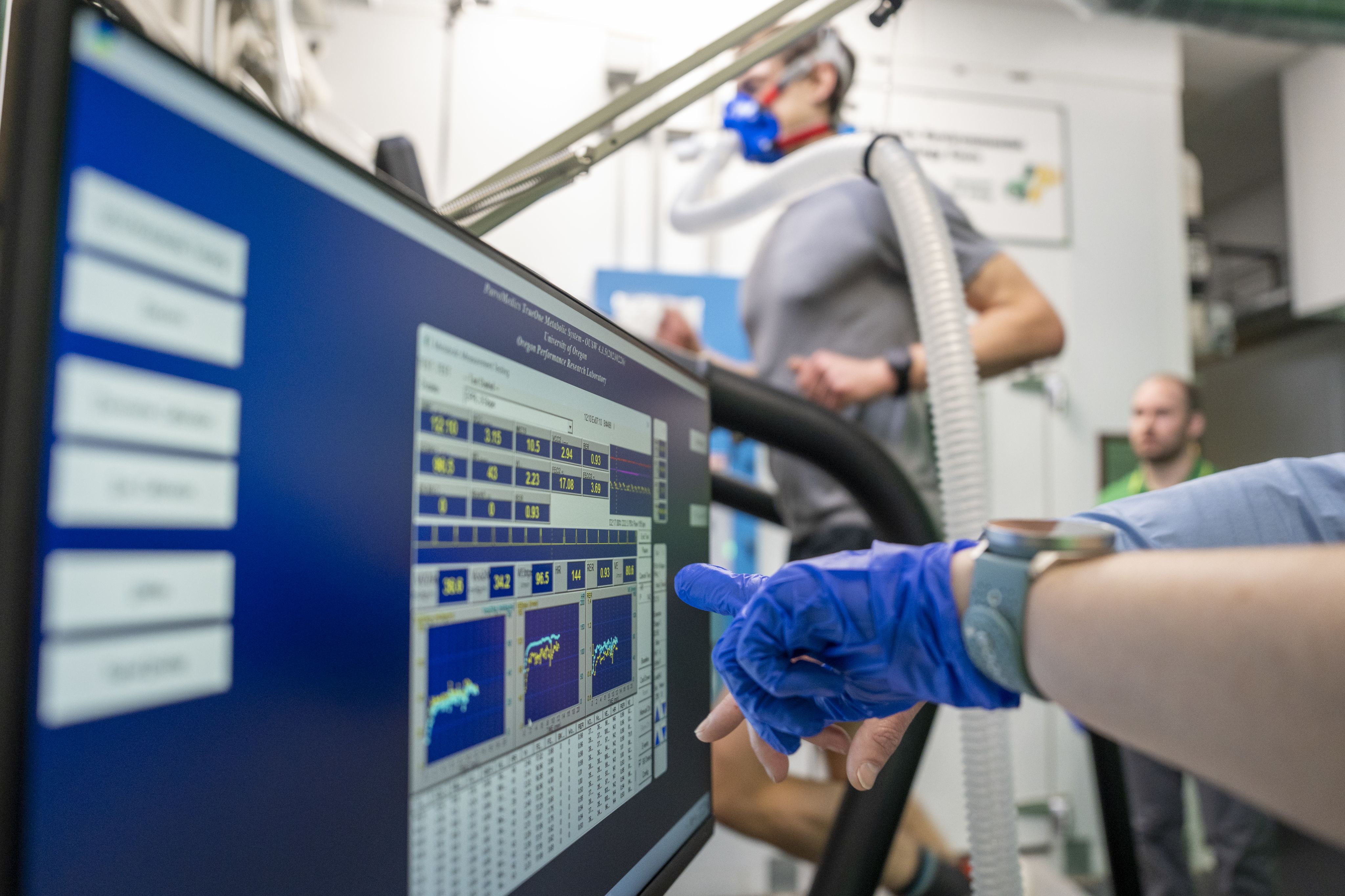 A gloved hand in the foreground pointing to data on a computer screen while an athlete in the background is running on a treadmill wearing a medical breathing apparatus.