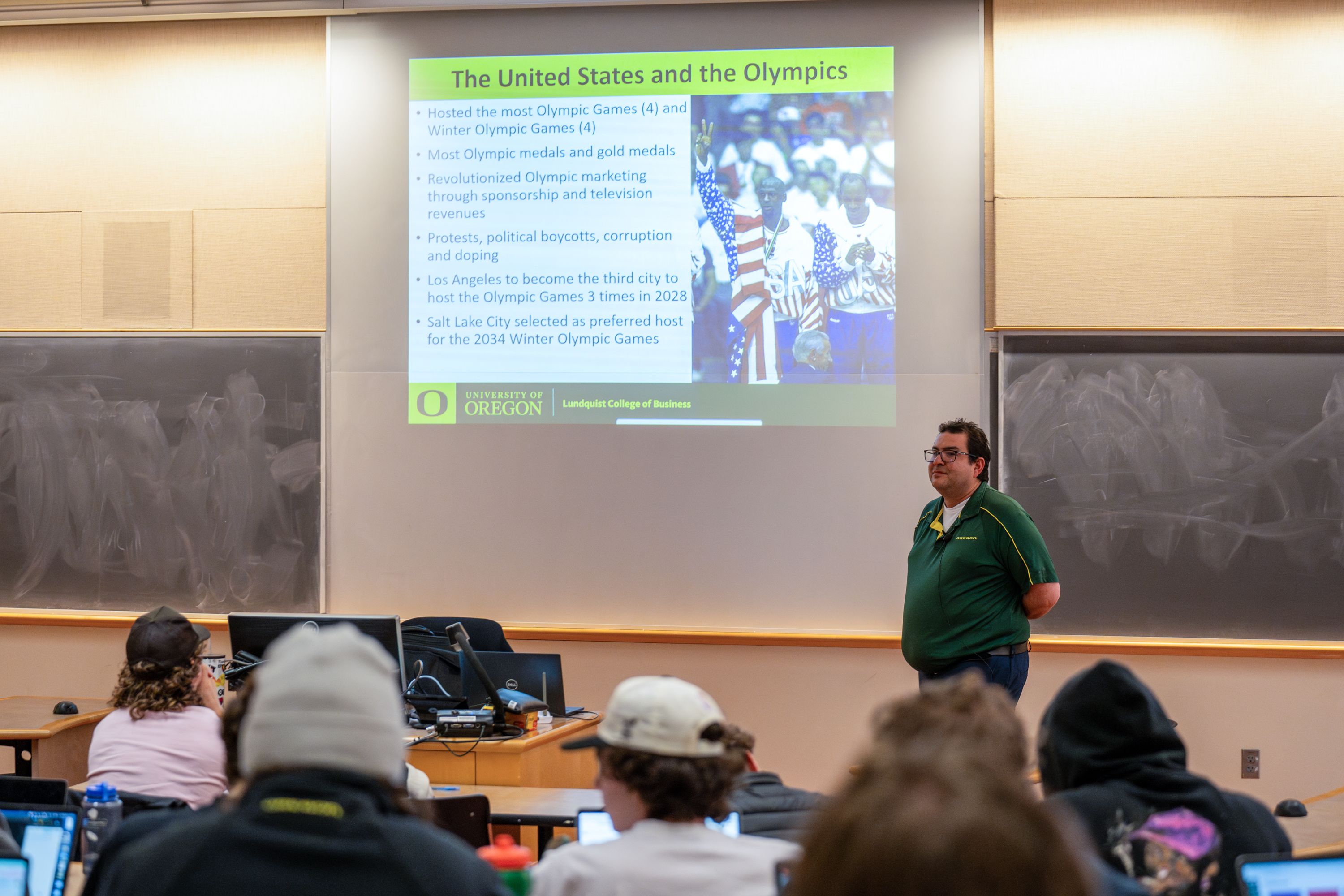 Yoav Dubinsky standing at the front of a classroom teaching with a presentation screen behind him showing a slide titled The United States and the Olympics.