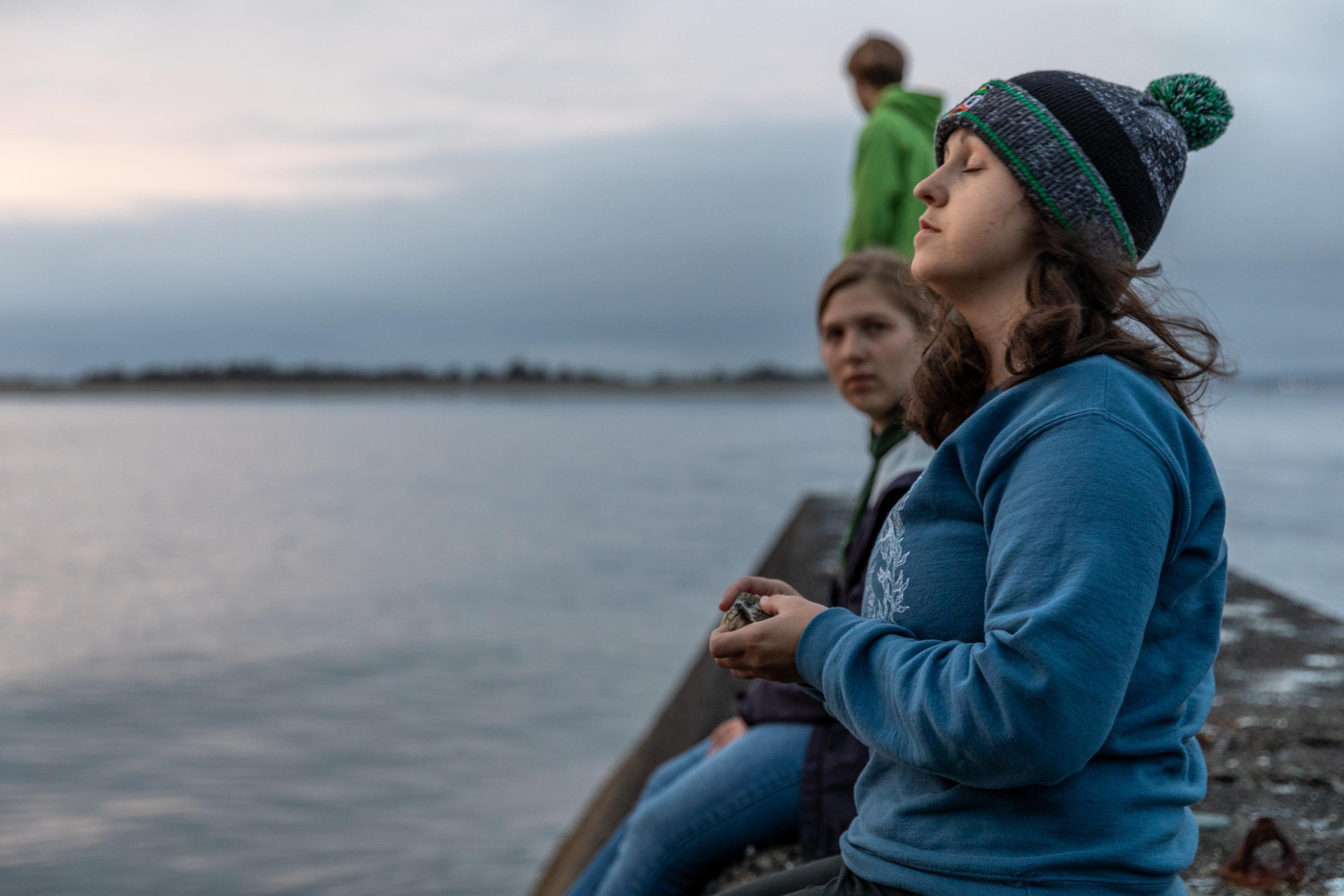 A student standing on a dock, faces the bay with eyes closed in meditation.